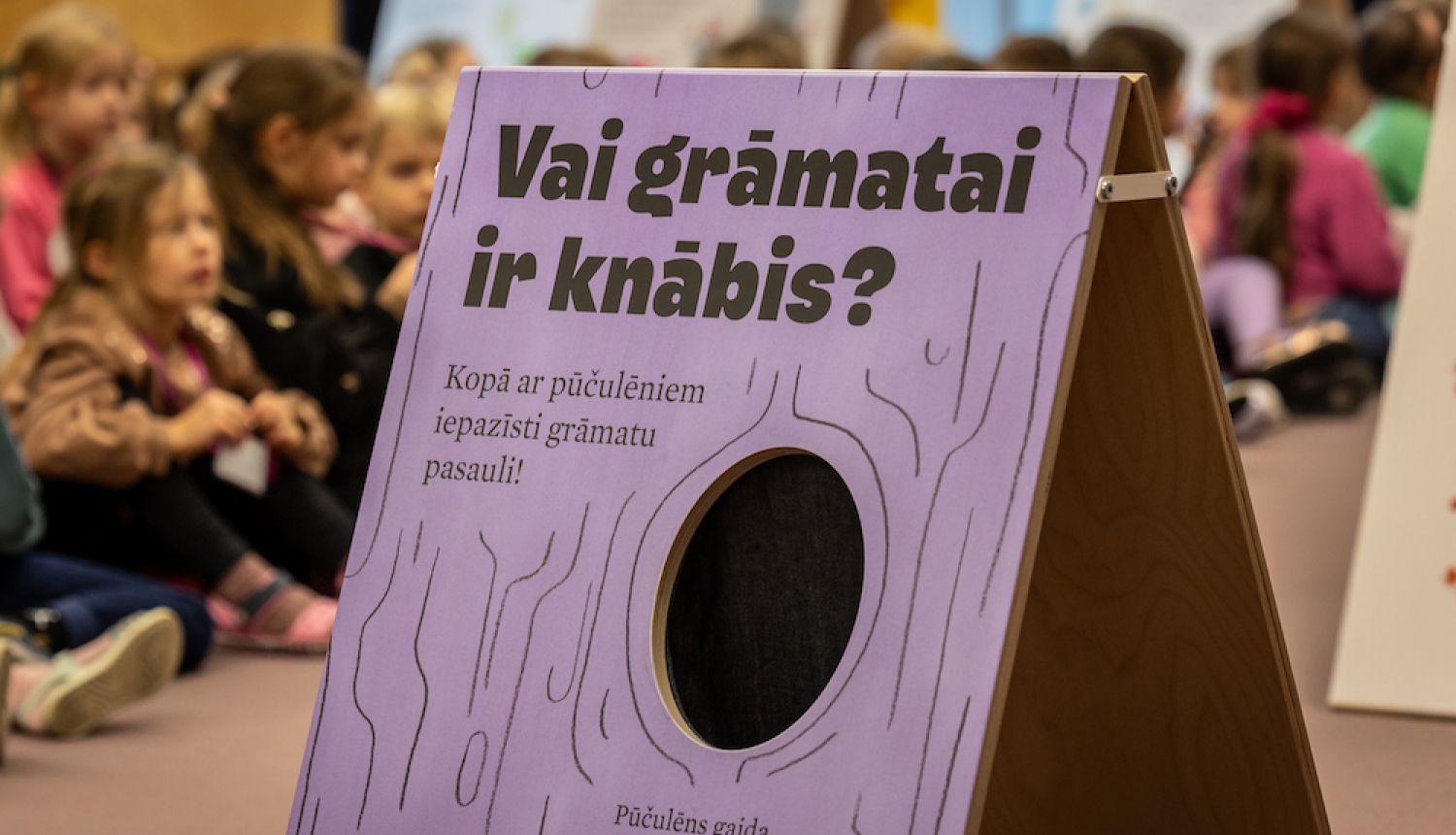 Bērni bibliotēkā un lillā baneris ar tekstu "Vai grāmai ir knābis" 